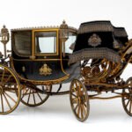 Eine schwarz-goldene verschnörkelte Pferdekutsche mit aufwändigen Details, großen Speichenrädern und dekorativen Laternen vor einem schlichten weißen Hintergrund.