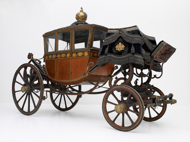 Verschnörkelte antike Pferdekutsche mit goldenen Details, geschlossener Kabine, großen Holzrädern und dekorativem schwarzem Verdeck auf weißem Hintergrund.