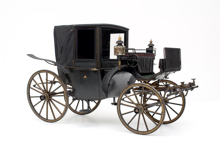 Eine schwarze, alte Pferdekutsche mit vier großen Holzrädern, einem überdachten Fahrgastraum und auf der Vorderseite angebrachten Laternen.