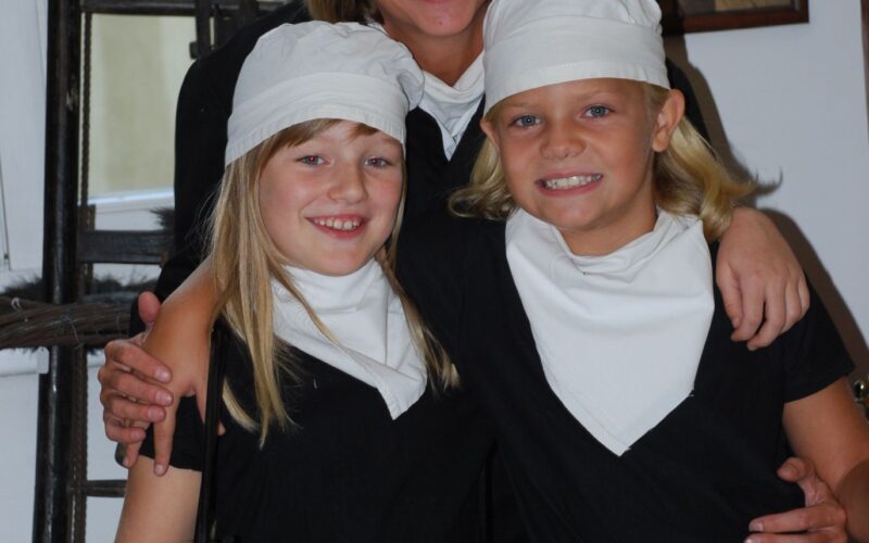 Eine Frau und zwei Kinder, alle schwarz gekleidet mit weißen Hüten und Schals, posieren in einem Haus. Ein Kind hält einen Schornsteinfegerpinsel.