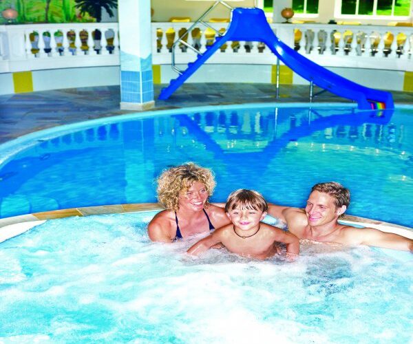 Eine Frau, ein Kind und ein Mann entspannen sich gemeinsam in einem runden Innen-Whirlpool neben einem Pool mit einer blauen Rutsche im Hintergrund.
