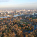 Luftaufnahme einer Stadtlandschaft mit einem Fluss, Parkanlagen, Baumgruppen und mehreren Hochhäusern unter klarem Himmel.