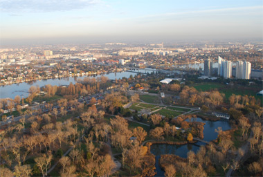 Luftaufnahme einer Stadtlandschaft mit einem Fluss, Parkanlagen, Baumgruppen und mehreren Hochhäusern unter klarem Himmel.