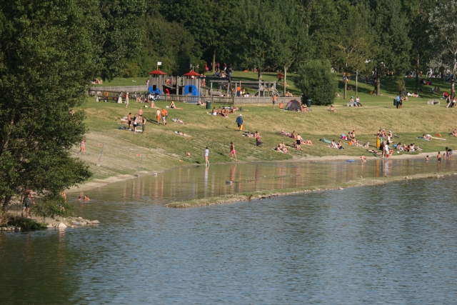 Menschen entspannen und sonnen sich auf einer Wiese am Seeufer in der Nähe eines Spielplatzes, mit Bäumen im Hintergrund und einigen Menschen, die im Wasser waten.