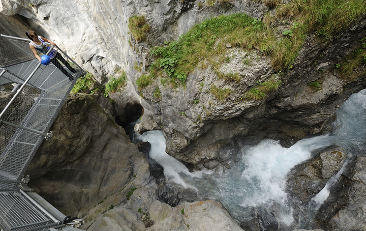 Eine Person steht auf einer Metallbrücke mit Blick auf eine felsige Schlucht mit schnell fließendem, türkisfarbenem Wasser darunter.