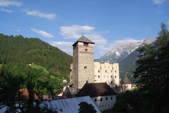 Eine Steinburg mit einem zentralen Turm, umgeben von Bäumen und Hügeln, mit Bergen im Hintergrund unter blauem Himmel.