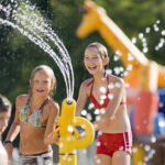Drei Kinder spielen mit einer Wasserspritzanlage in einem Wasserpark im Freien, wobei im Hintergrund aufblasbare Strukturen und Wassertropfen zu sehen sind.