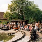 Eine Menschenmenge, darunter viele Kinder, sitzt und steht um eine Miniatureisenbahnstrecke in der Nähe eines Teiches, mit einem kleinen Zug und Häusern im Hintergrund.