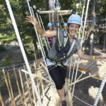 Eine Person, die eine Sicherheitsausrüstung trägt, lächelt und balanciert auf Seilen in einem Outdoor-Abenteuerpark zwischen Bäumen.