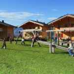 Kinder spielen auf einer Wippe und einem Spielplatz vor drei Holzhäusern im Chalet-Stil auf einer Wiese mit Bergen im Hintergrund.