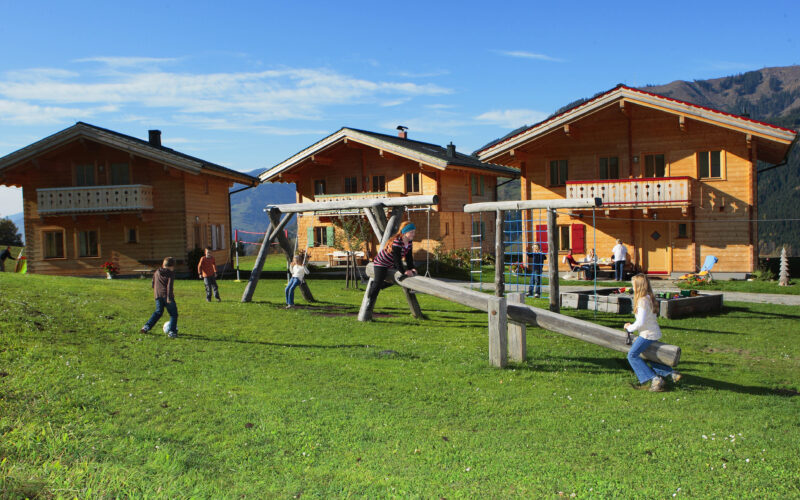 Kinder spielen auf einer Wippe und einem Spielplatz vor drei Holzhäusern im Chalet-Stil auf einer Wiese mit Bergen im Hintergrund.