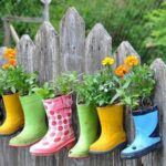 Bunte, mit Blumen gefüllte Gummistiefel hängen an einem Holzzaun und dienen als kreative Pflanzgefäße.