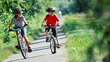 Zwei Kinder mit Helmen fahren mit dem Fahrrad auf einem gepflasterten Weg im Grünen.