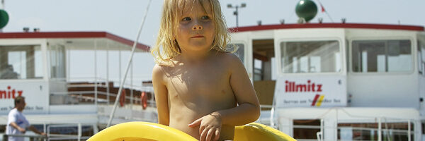 Ein kleines Kind mit langen Haaren steht mit einem gelben aufblasbaren Ring in der Hand vor angedockten Booten und einem sonnigen Himmel im Hintergrund.