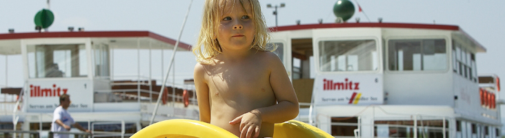 Ein kleines Kind mit langen Haaren steht mit einem gelben aufblasbaren Ring in der Hand vor angedockten Booten und einem sonnigen Himmel im Hintergrund.