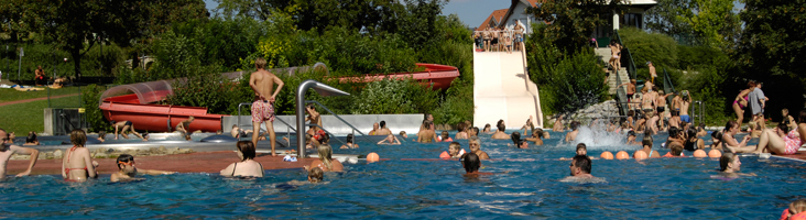 Freibad mit vielen Menschen; einige schwimmen, andere sind in der Nähe der Rutschen und auf dem Pooldeck versammelt. Im Hintergrund sind Bäume und Gebäude zu sehen.