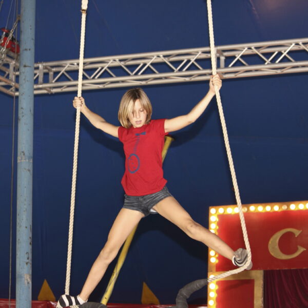 Ein Kind in rotem Hemd und kurzer Hose balanciert auf einem Zirkusseilgerät und hält sich an zwei Seilen unter einem großen blauen Zirkuszelt fest.