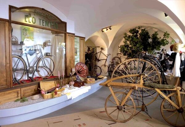 Eine Museumsausstellung mit antiken Fahrrädern, einem hölzernen Dreirad, alten Gegenständen und dekorativen Pflanzen unter Gewölbedecken.