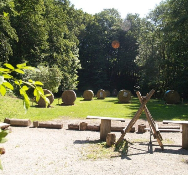 Ein Bogenschießplatz im Freien mit Holzbänken im Vordergrund und mehreren großen Strohzielen, die auf einer von Bäumen umgebenen Wiese aufgestellt sind.