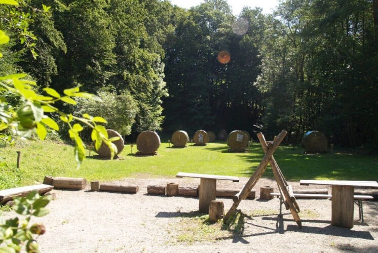 Ein Bogenschießplatz im Freien mit Holzbänken im Vordergrund und mehreren großen Strohzielen, die auf einer von Bäumen umgebenen Wiese aufgestellt sind.