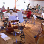 Mehrere Musiker in einem Proberaum mit verstreuten Stühlen, Notenständern, Notenblättern und Streichinstrumenten, darunter Bässe und Geigen.