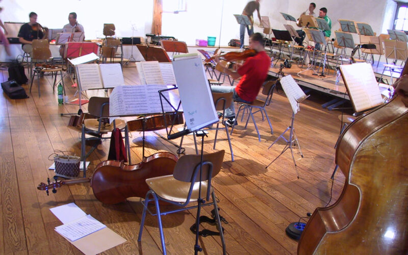 Mehrere Musiker in einem Proberaum mit verstreuten Stühlen, Notenständern, Notenblättern und Streichinstrumenten, darunter Bässe und Geigen.