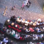 Ein Künstler steht vor einem sitzenden Publikum, das in Reihen auf einem kopfsteingepflasterten Innenhof angeordnet ist, von oben gesehen.