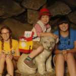 Drei Kinder sitzen in einer Steinhöhle; eines sitzt auf einer weißen Bärenstatue, während die beiden anderen an den Seiten sitzen und in die Kamera lächeln.