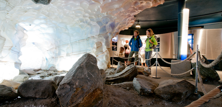 Zwei Personen beobachten eine Indoor-Gletscherausstellung mit simulierten Eiswänden, großen Felsen und Informationstafeln hinter einer Seilbarriere.