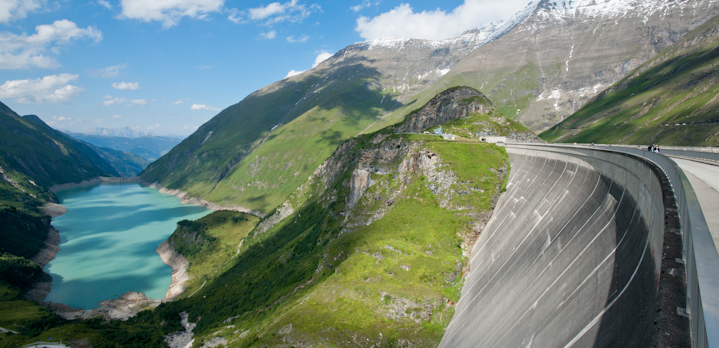 Ein großer geschwungener Staudamm staut türkisfarbenes Wasser in einem Bergtal, umgeben von steilen, grünen Hängen und teilweise schneebedeckten Gipfeln unter blauem Himmel.