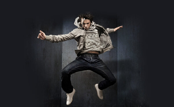 Eine Person in legerer Kleidung und weißen Turnschuhen befindet sich in der Luft und springt energisch vor einem dunklen, minimalistischen Hintergrund.