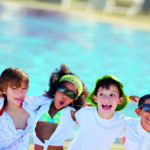 Vier Kinder in Sommerkleidung und mit Sonnenbrille stehen lächelnd und grinsend vor einem Schwimmbad.