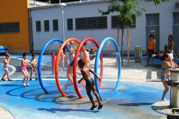 Kinder in Badeanzügen spielen an einem sonnigen Tag in einem Wasserpark und rennen durch bunte, runde Sprinkler, während Erwachsene in der Nähe zuschauen.