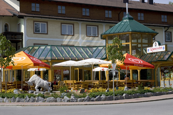 Außensitzbereich eines Restaurants mit bunten Sonnenschirmen, einem glasüberdachten Bereich und einer dekorativen Stierstatue vor einem mehrstöckigen Gebäude.