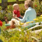 Ein Erwachsener und ein Baby sitzen zusammen auf einem Liegestuhl in einem Garten, einander zugewandt und lächelnd, mit Blumen und einem Holzzaun im Hintergrund.
