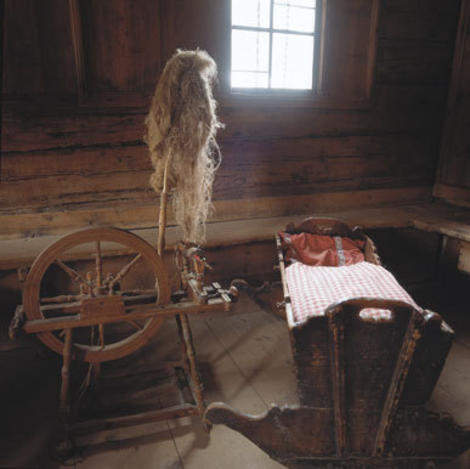 In einem rustikalen, sonnendurchfluteten Holzraum stehen ein Spinnrad mit Flachs und eine Holzwiege mit einer rotkarierten Decke.