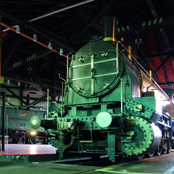 Eine alte Dampflokomotive mit der Nummer 1800 steht in einem schwach beleuchteten Lokschuppen mit Industriebalken und roten Dachziegeln darüber.