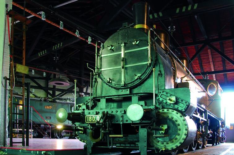 Eine alte Dampflokomotive mit der Nummer 1800 steht in einem schwach beleuchteten Lokschuppen mit Industriebalken und roten Dachziegeln darüber.