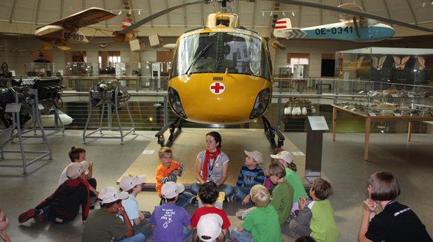 Eine Gruppe Kinder sitzt vor einem gelben Rettungshubschrauber in einem Museum und hört einer Frau zu.