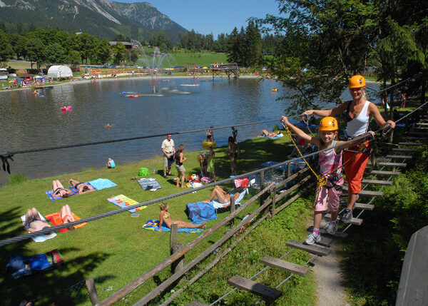 Menschen entspannen am Seeufer, während im Vordergrund eine Frau und ein Kind mit Helmen einen Seilparcours absolvieren. Im Hintergrund sind Bäume, Gras und Berge zu sehen.