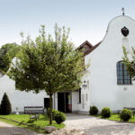 Ein weißes, historisches Gebäude mit Rundbogenfenstern und einem geschwungenen Giebel, umgeben von Bäumen, Bänken und einem Kopfsteinpflasterweg, der zum Eingang führt.