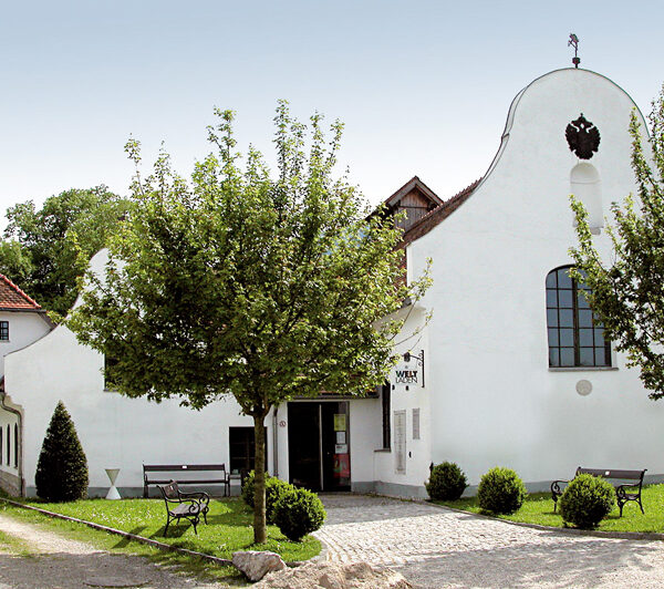 Ein weißes, historisches Gebäude mit Rundbogenfenstern und einem geschwungenen Giebel, umgeben von Bäumen, Bänken und einem Kopfsteinpflasterweg, der zum Eingang führt.