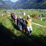 Eine Gruppe von Kindern, angeführt von einem Erwachsenen, geht in einer Reihe auf einen grasbewachsenen Hügel mit einem Dorf und Bergen im Hintergrund.