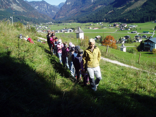 Eine Gruppe von Kindern, angeführt von einem Erwachsenen, geht in einer Reihe auf einen grasbewachsenen Hügel mit einem Dorf und Bergen im Hintergrund.