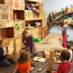 Kinder spielen mit Holzklötzen und Kisten in einem Raum mit einem baumförmigen Bücherregal, Büchern und Kissen. Einige Kinder bauen, andere erkunden die Umgebung oder sitzen auf einem großen Sitzsack.