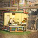 Bunt getäfelter Raum mit großem Fenster, von außen betrachtet; drinnen sitzt eine Person an einem Tisch mit Malutensilien. Der Raum ist von künstlichen Felsen und Kletternetzen umgeben.