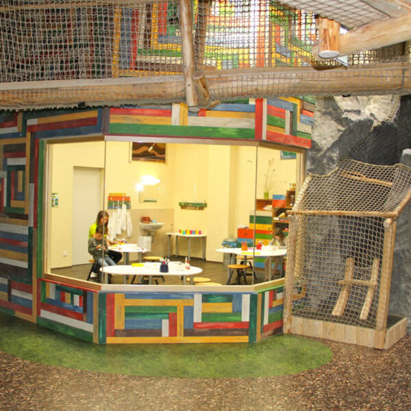 Bunt getäfelter Raum mit großem Fenster, von außen betrachtet; drinnen sitzt eine Person an einem Tisch mit Malutensilien. Der Raum ist von künstlichen Felsen und Kletternetzen umgeben.