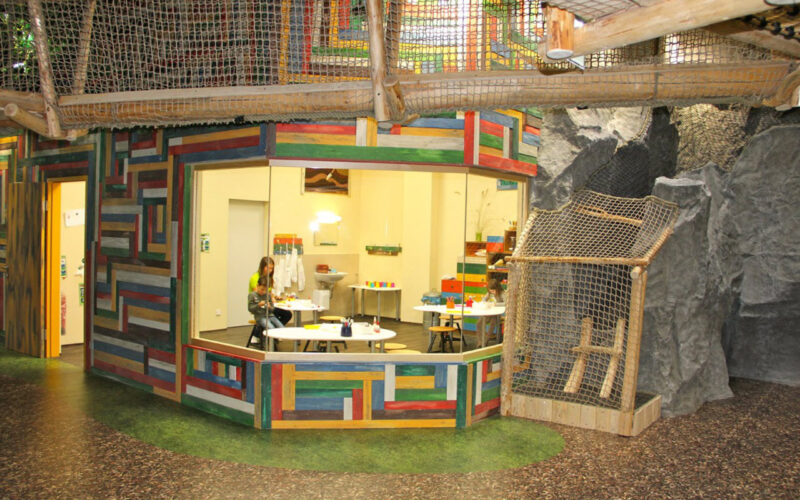 Bunt getäfelter Raum mit großem Fenster, von außen betrachtet; drinnen sitzt eine Person an einem Tisch mit Malutensilien. Der Raum ist von künstlichen Felsen und Kletternetzen umgeben.