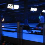 Eine Person bereitet sich darauf vor, in einer schwach beleuchteten Bowlingbahn mit leuchtenden Bahnen und Bildschirmen über der Decke zu bowlen.