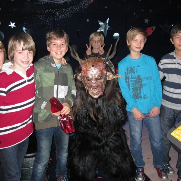 Eine Gruppe von fünf Jungen steht in einem Raum mit Sternenhimmel im Hintergrund um eine Person im Kostüm eines gehörnten Monsters herum; ein Junge macht ein Peace-Zeichen.